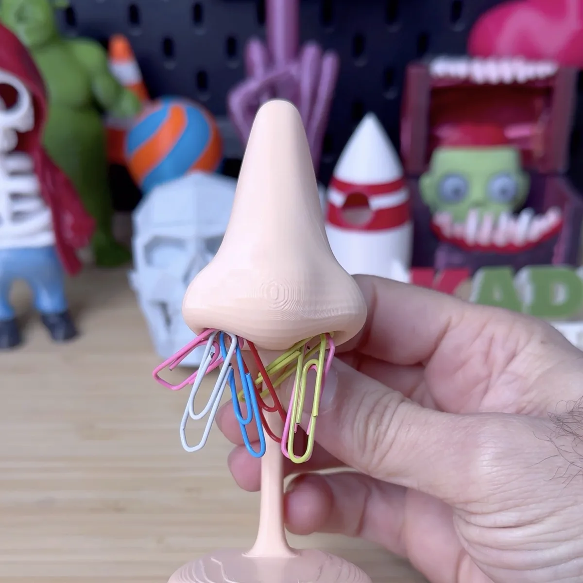 A hand holds a 3D-printed, flesh-toned nose-shaped object with multiple colorful paperclips stuck into its nostril area. The…