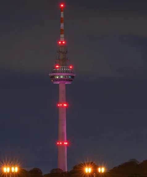 A tall, slender tower with red lights at various levels stands against a dark night sky, with streetlights glowing in the…