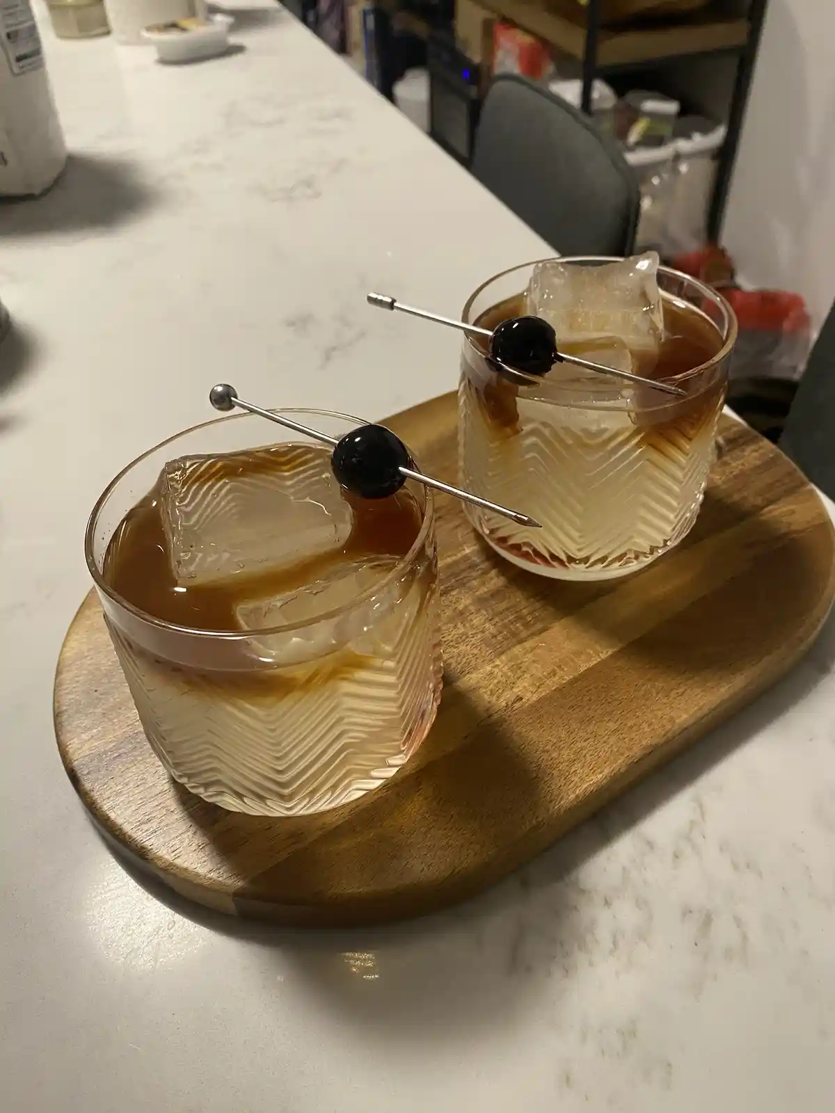 Two identical glasses of amber-colored cocktail with large ice cubes and a single maraschino cherry on a metal pick, resting…
