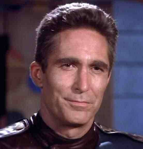 A close-up of actor James Brolin as a character in a dark uniform, smiling subtly with a confident, slightly smug…
