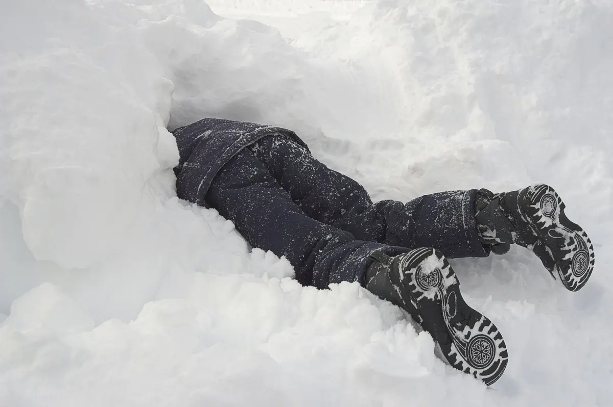 a person is lying face-down in a deep pile of snow, with only their legs, dark snow-speckled pants, and black winter boots…