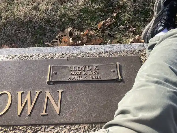 A close-up photo shows a person's leg and sneaker resting on a gravestone. The stone has a bronze plaque engraved with…
