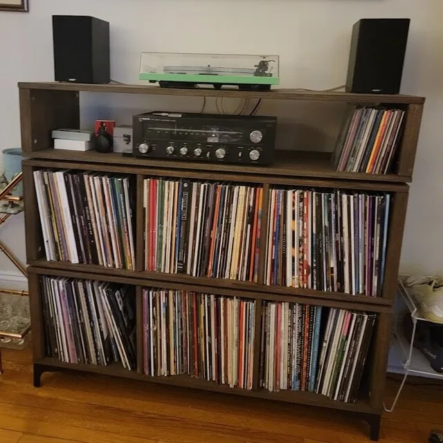 A wooden shelving unit filled with vinyl records on all shelves, topped with a turntable and two black speakers. The text 'i…