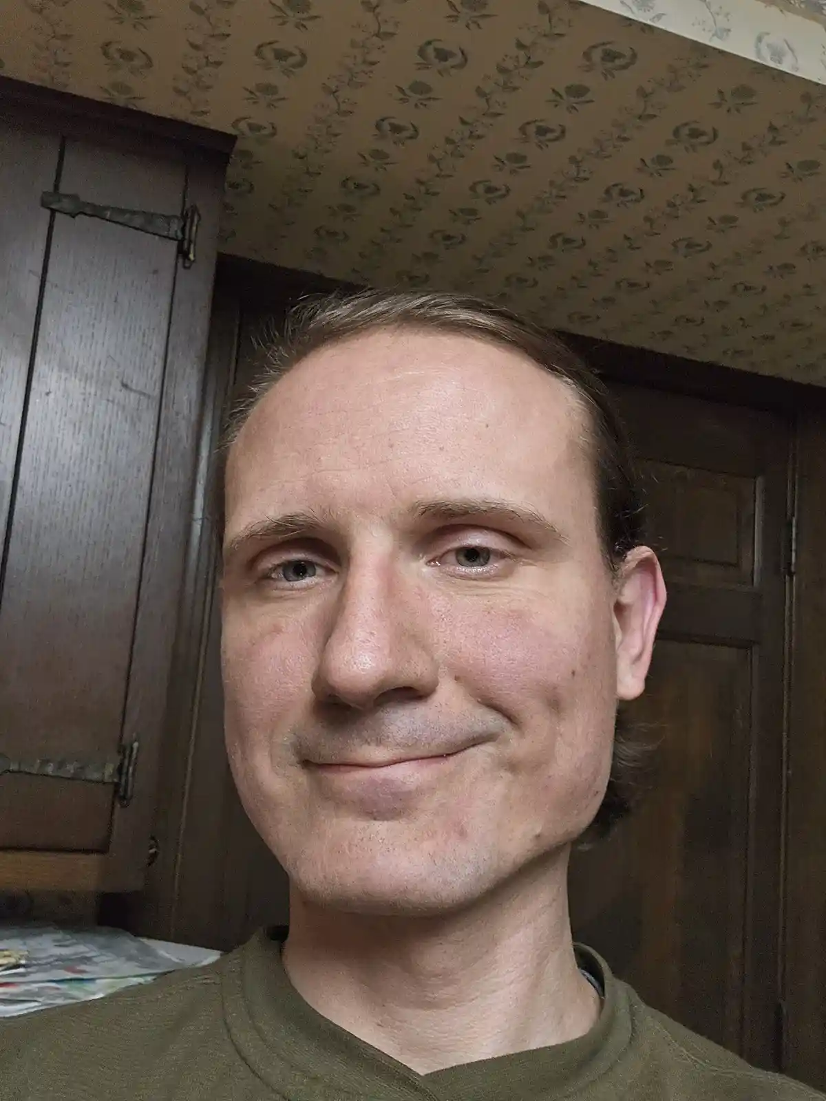 A man with a subtle, slightly smug smile looks at the camera, standing in front of dark wooden doors and patterned…