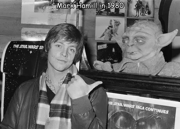 A black-and-white photo of young Mark Hamill in 1980, wearing a jacket and scarf, pointing at a Yoda head prop beside him…