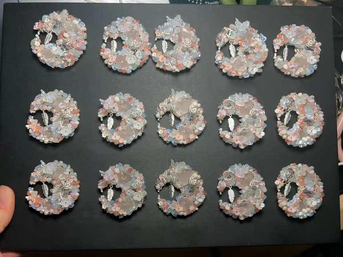 A hand holds a black display board showcasing fifteen identical circular brooches, each intricately crafted with pink, blue…