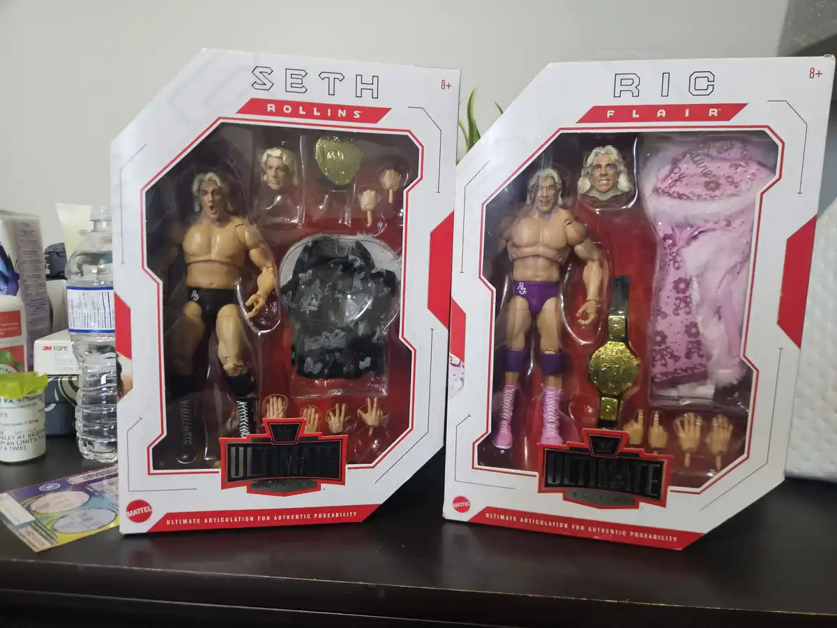 Two WWE Ultimate Edition action figure boxes sit side by side on a dark surface, one labeled 'Seth Rollins' with black…