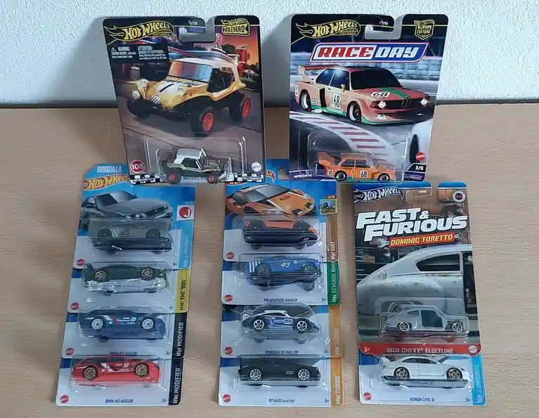 Several Hot Wheels toy car packages are arranged on a wooden surface, featuring various models including a yellow dune…