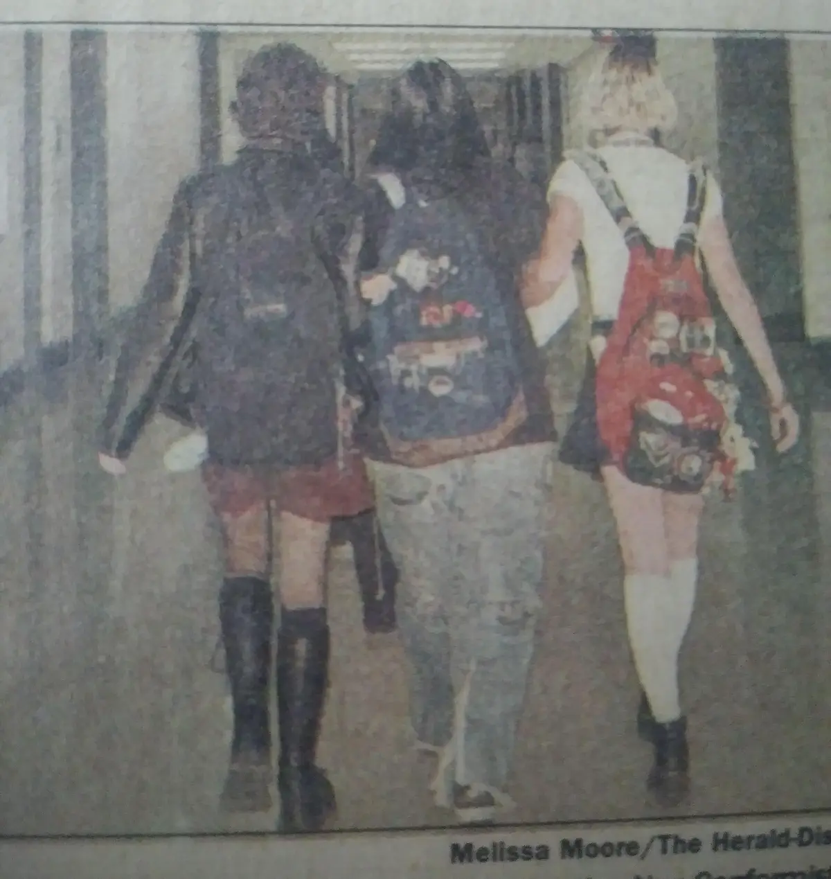 Three young women walk away from the camera down a hallway, each wearing a heavily decorated backpack. The woman on the left…