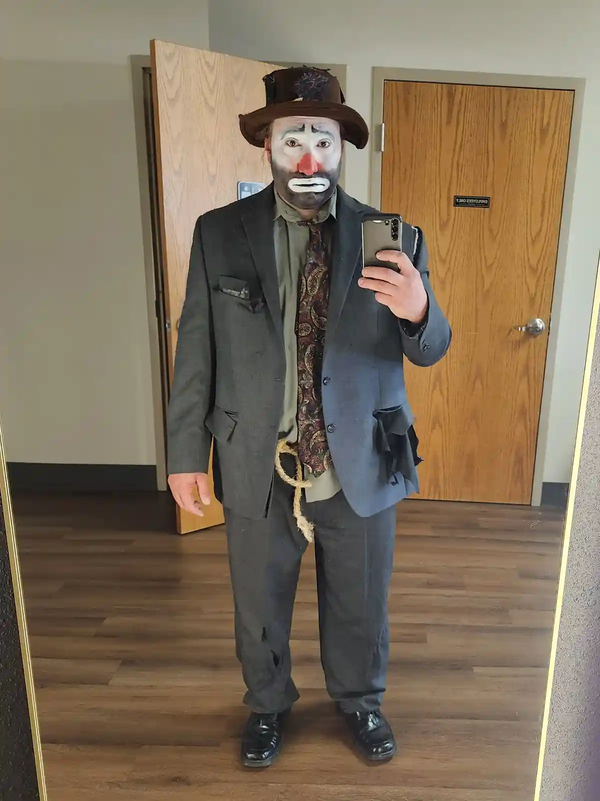 A person in a mirror selfie wearing a tattered suit, a paisley tie, and a clown makeup with a red nose and white face paint…