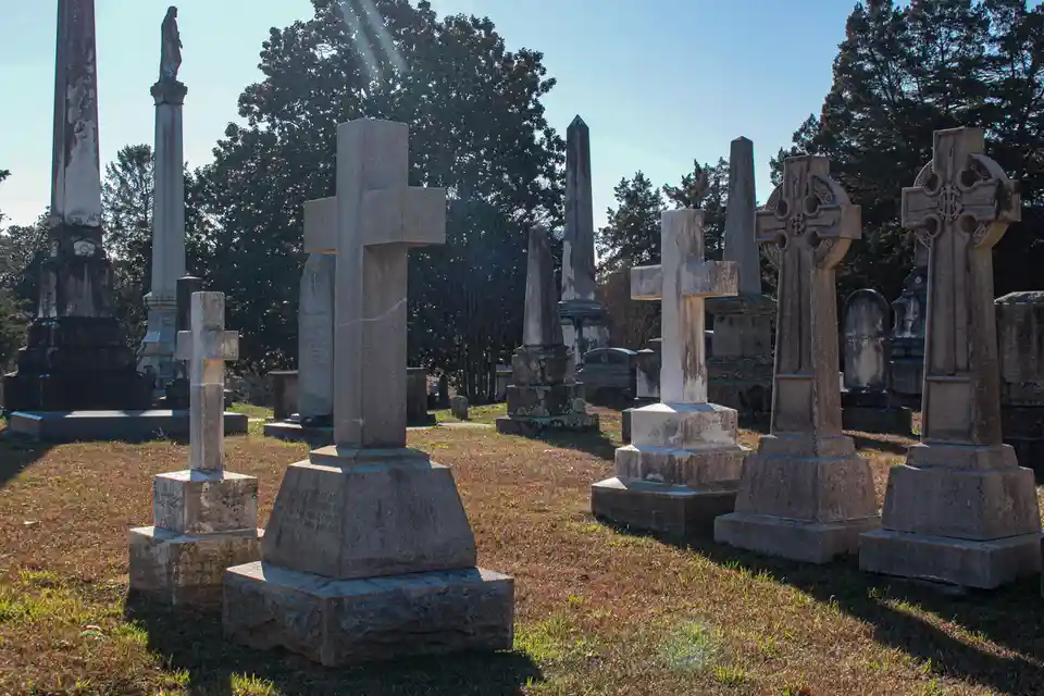 A sunlit cemetery scene with numerous weathered tombstones, crosses, and obelisks standing in grassy rows under a clear blue…