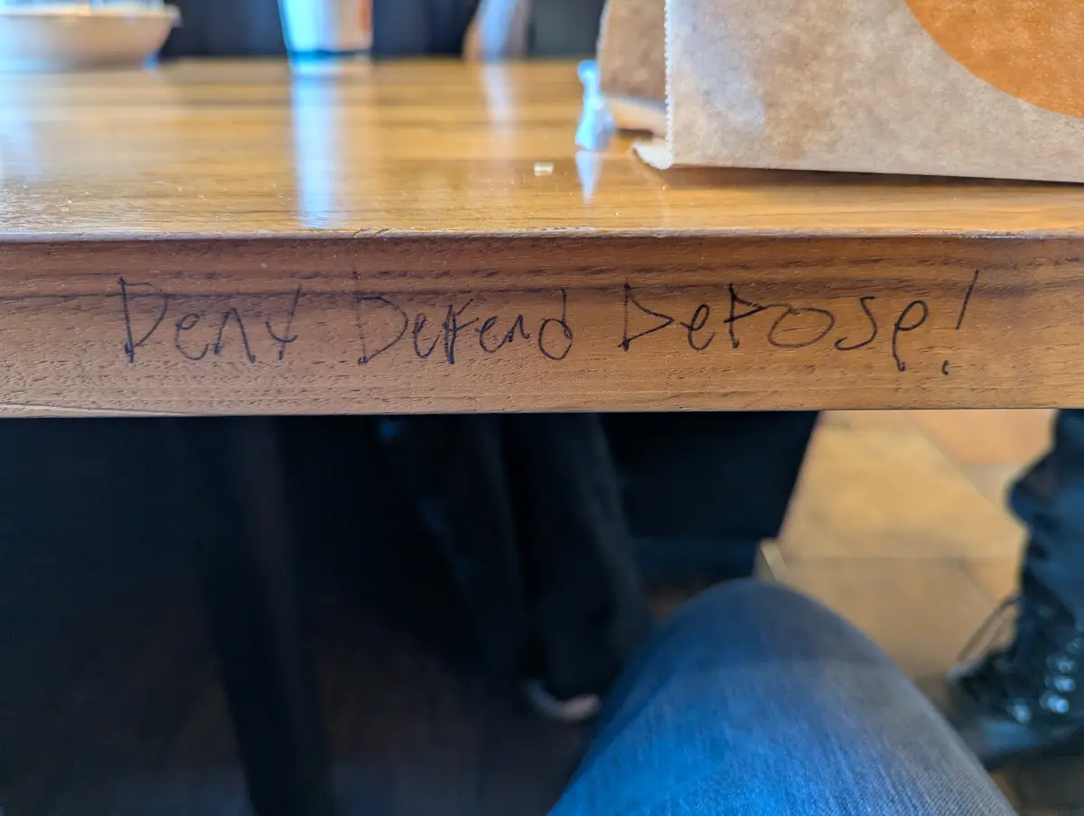 A close-up photo shows the edge of a wooden table with the handwritten phrase 'Deny Defend Depose!' scratched into the wood…