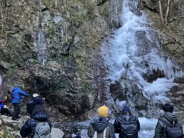 a group of people in winter jackets stand before a rocky cliff face where a waterfall is partially frozen into thick icicles…