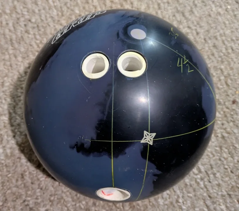 A close-up photo of a dark blue bowling ball resting on carpet, showing its three finger holes, a star-shaped sticker, and…