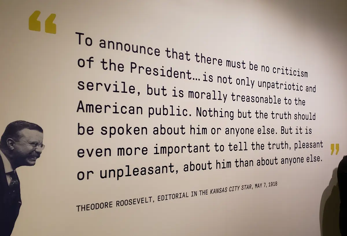 A wall display features a black-and-white portrait of Theodore Roosevelt on the left, with a large block of text quoting…