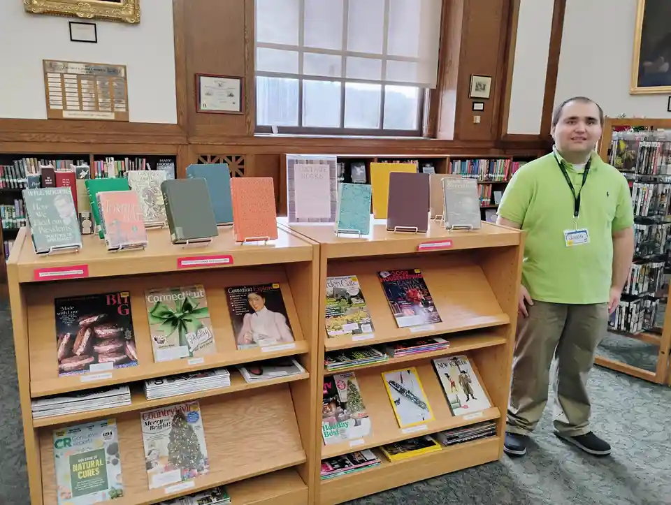 A man in a bright green polo shirt and khaki pants stands beside a wooden magazine and book display in a library. He wears a…