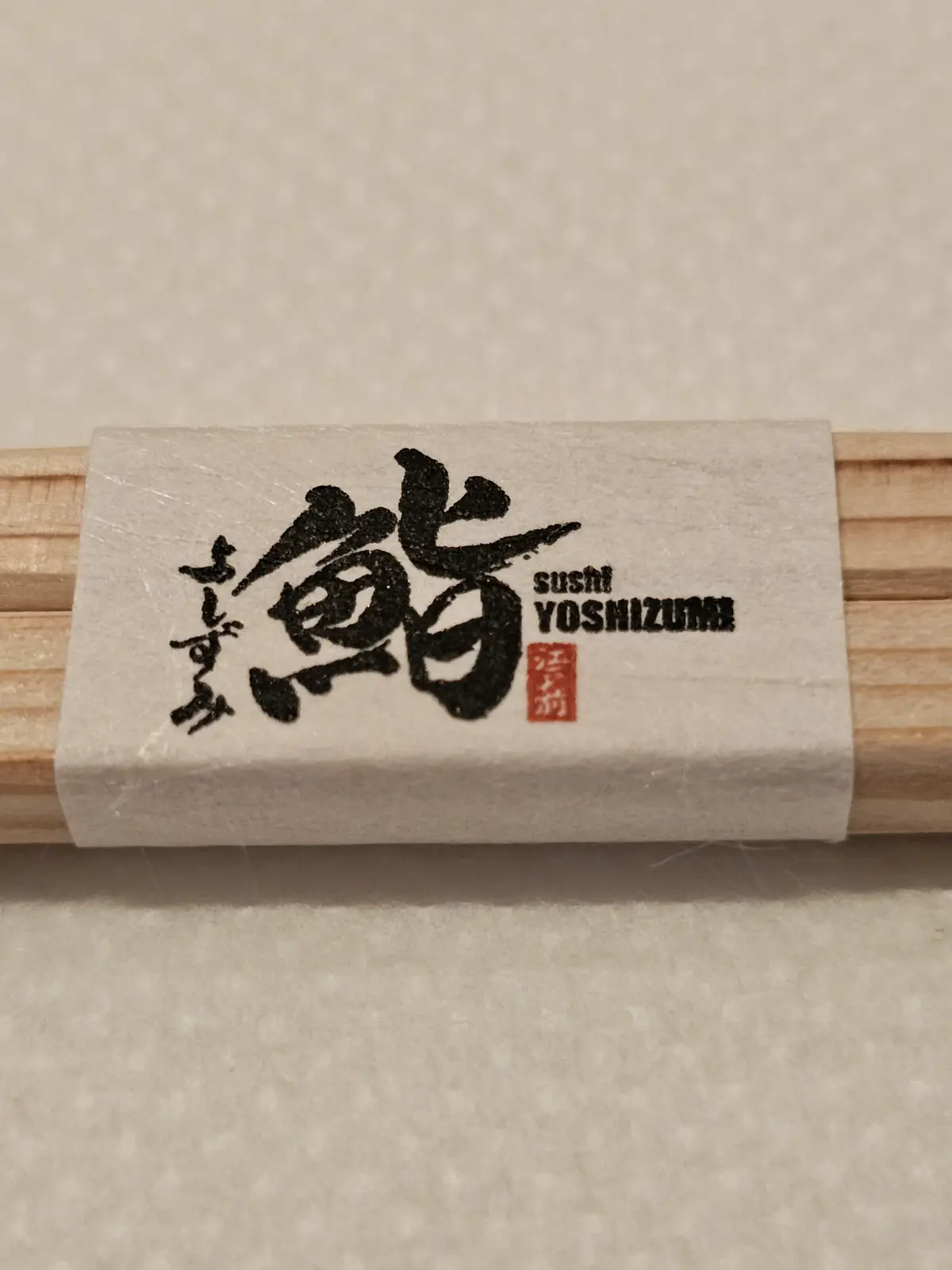 A close-up of wooden chopsticks wrapped with a white paper sleeve featuring large black Japanese calligraphy, the text…