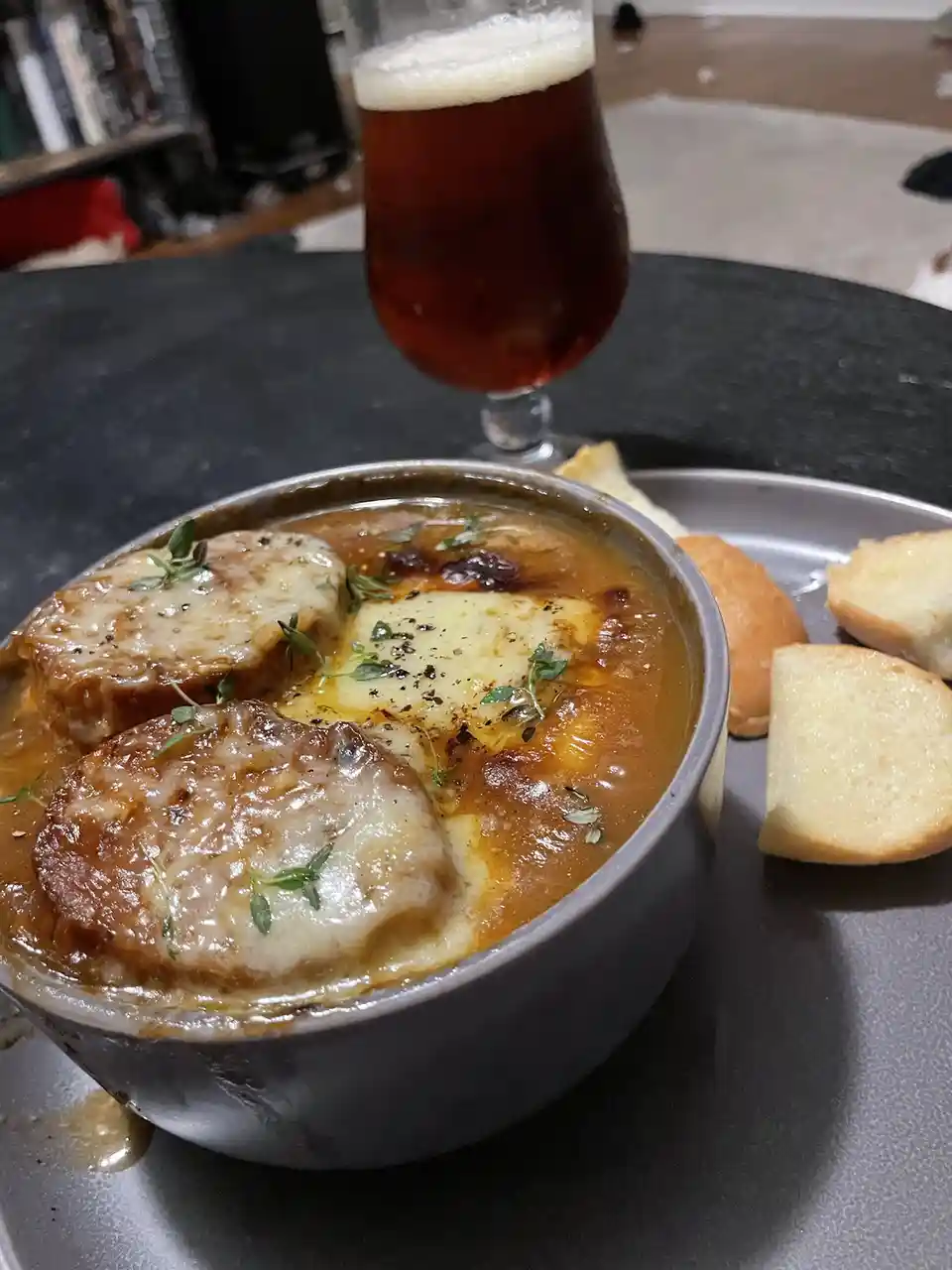 A close-up photo shows a bowl of French onion soup with melted cheese and herbs, accompanied by sliced bread and a glass of…