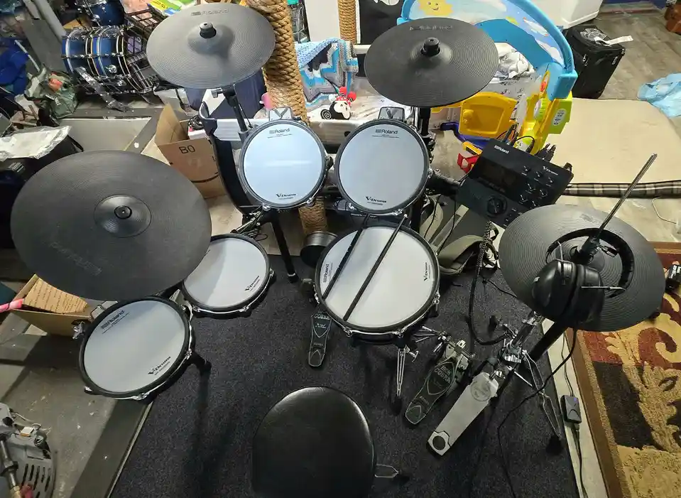 An overhead shot of a Roland electronic drum set in a cluttered room, with a pair of headphones hanging on a cymbal and a…
