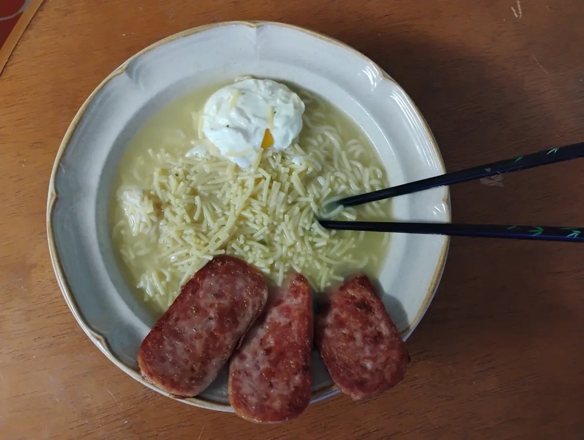 A bowl of instant noodles with a poached egg on top, three slices of sausage beside it, and chopsticks resting in the…