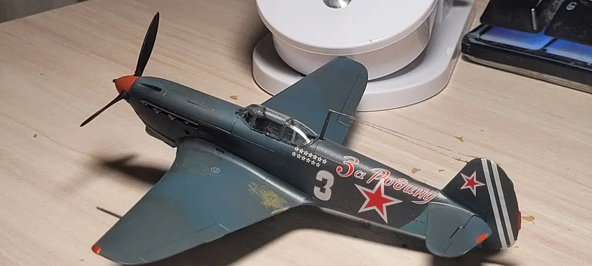 A detailed model of a Soviet fighter plane, likely a Yak-1, rests on a wooden desk. It features dark green paint, red stars…