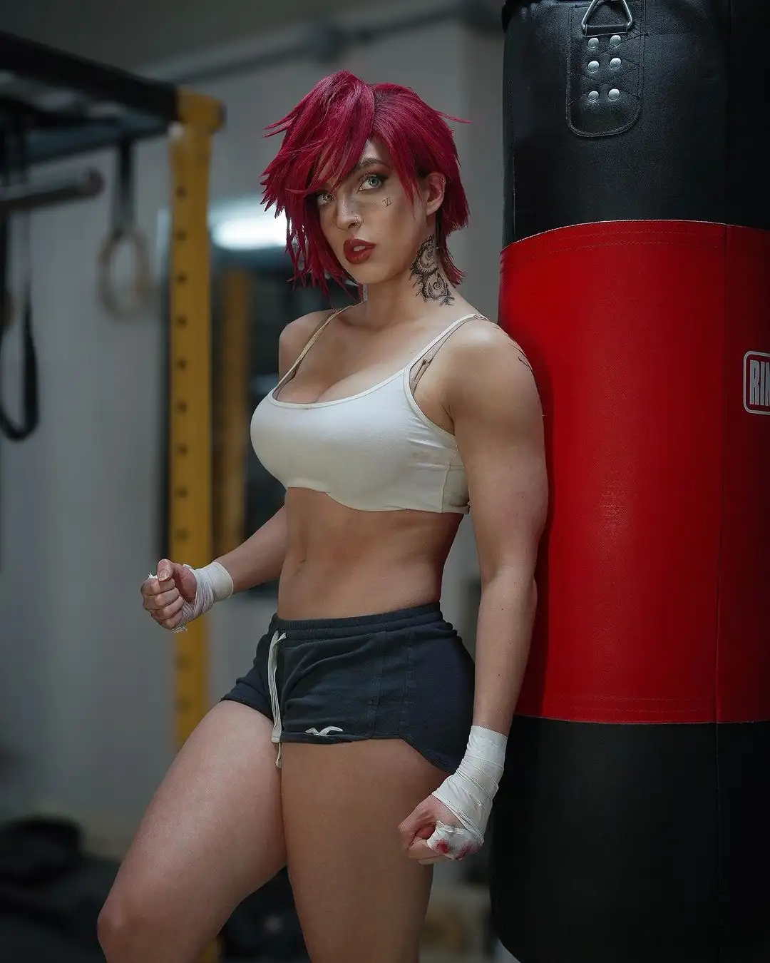 A woman with short red hair and a visible tattoo on her neck stands in a gym next to a red and black punching bag, wearing a…