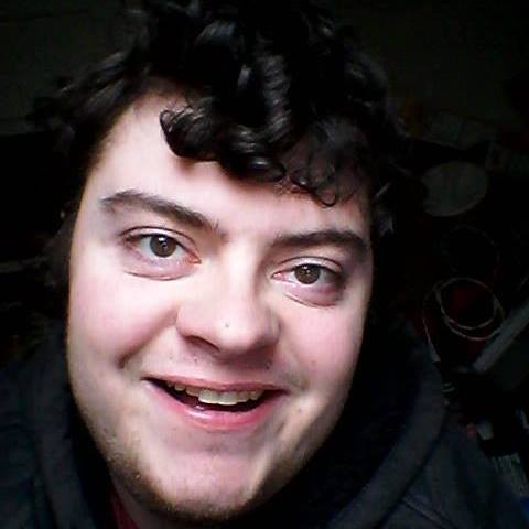 A close-up selfie of a young man with dark curly hair and a wide, toothy grin, looking directly at the camera. He has light…