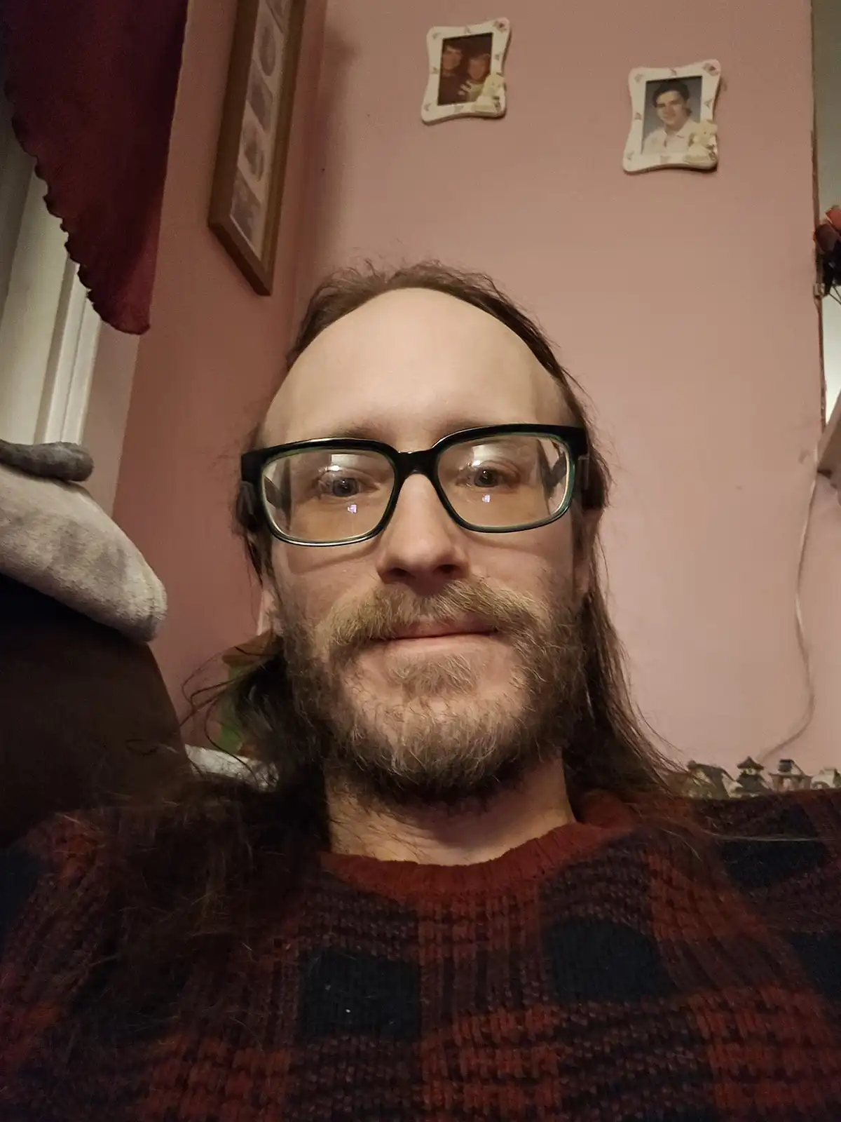 A man with long hair, a beard, and glasses looks at the camera with a slight smile while wearing a red and black plaid…