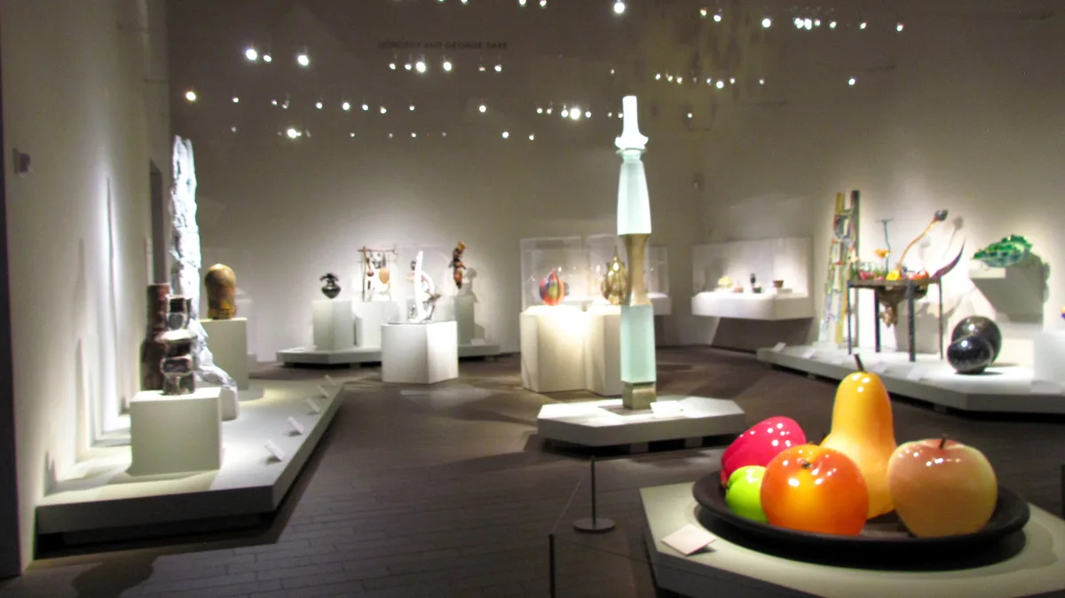 A museum gallery filled with various glass sculptures on white pedestals under bright spotlights, with a plate of colorful…