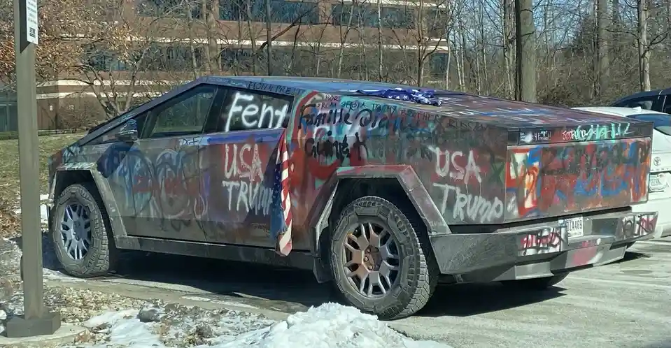 A heavily graffiti-covered Tesla Cybertruck parked in a snowy lot, with visible text including 'Fent', 'USA TRUMP', and…