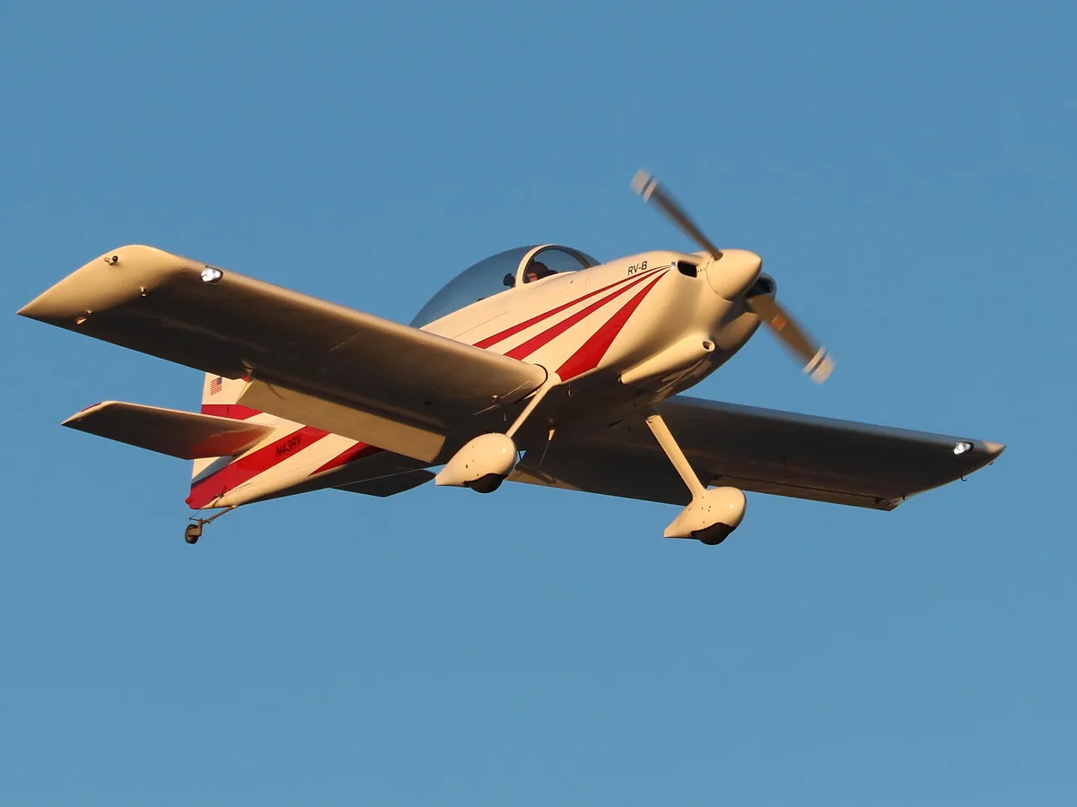 A white and red RV-8 single-engine airplane with registration N43RV is flying against a clear blue sky, its propeller…