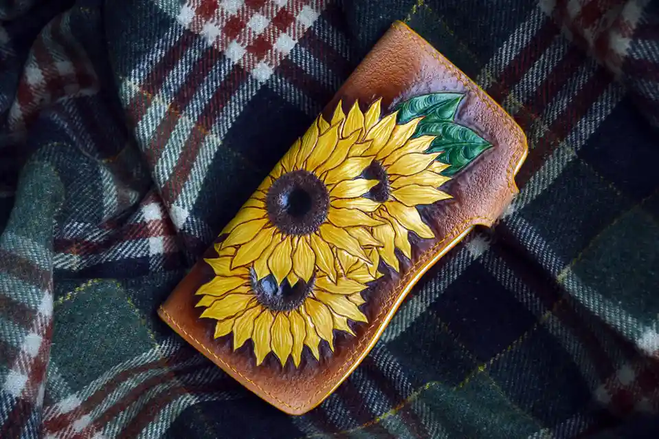 A brown leather wallet with intricate hand-painted sunflowers and green leaves rests on a plaid blanket with red, white, and…
