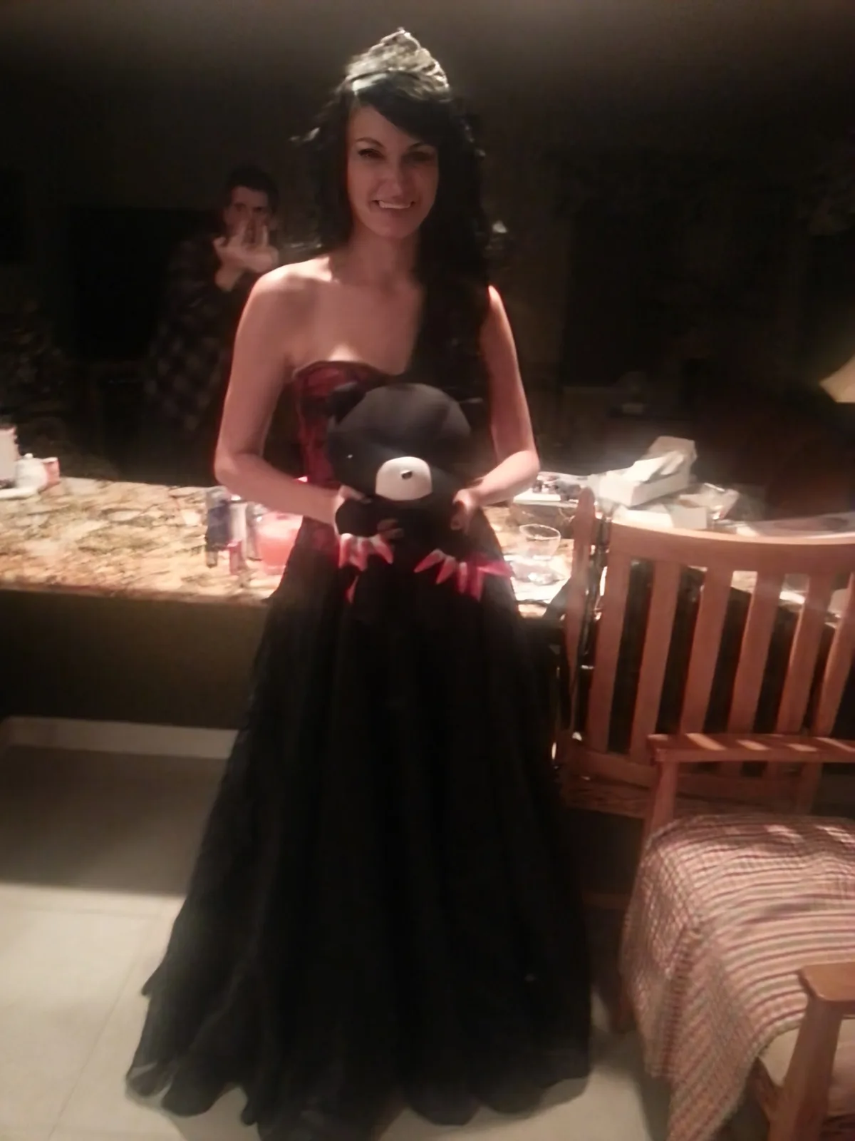 A woman in a black and red formal gown with a tiara smiles while holding a black plush toy with a white face and red fangs…