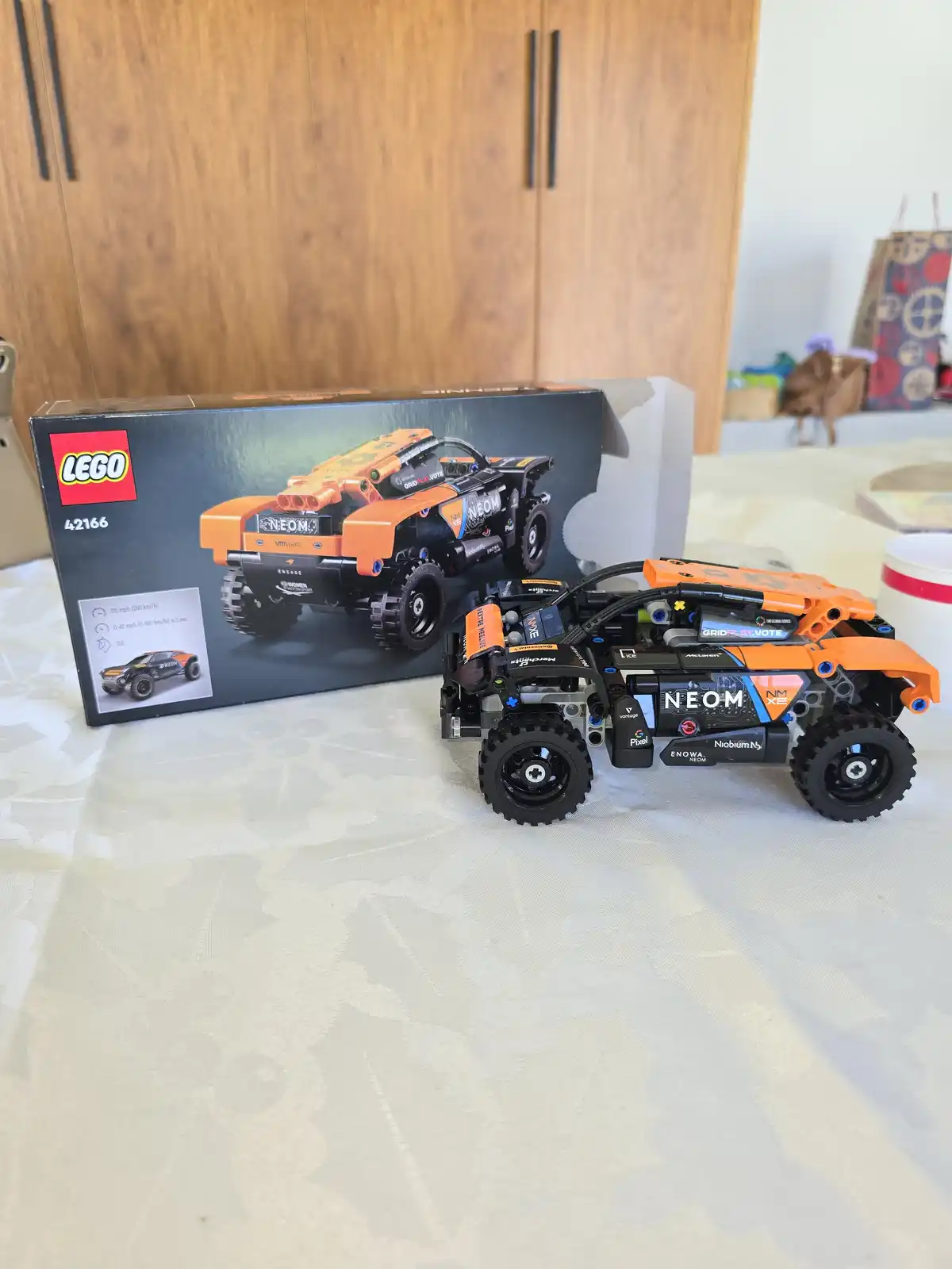 A completed orange and black LEGO Technic 42166 NEOM off-road vehicle sits on a patterned tablecloth next to its original…