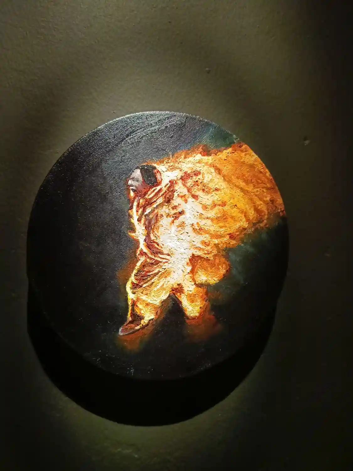 A circular painting depicts a figure in a spacesuit engulfed in flames against a dark background, illuminated by a…