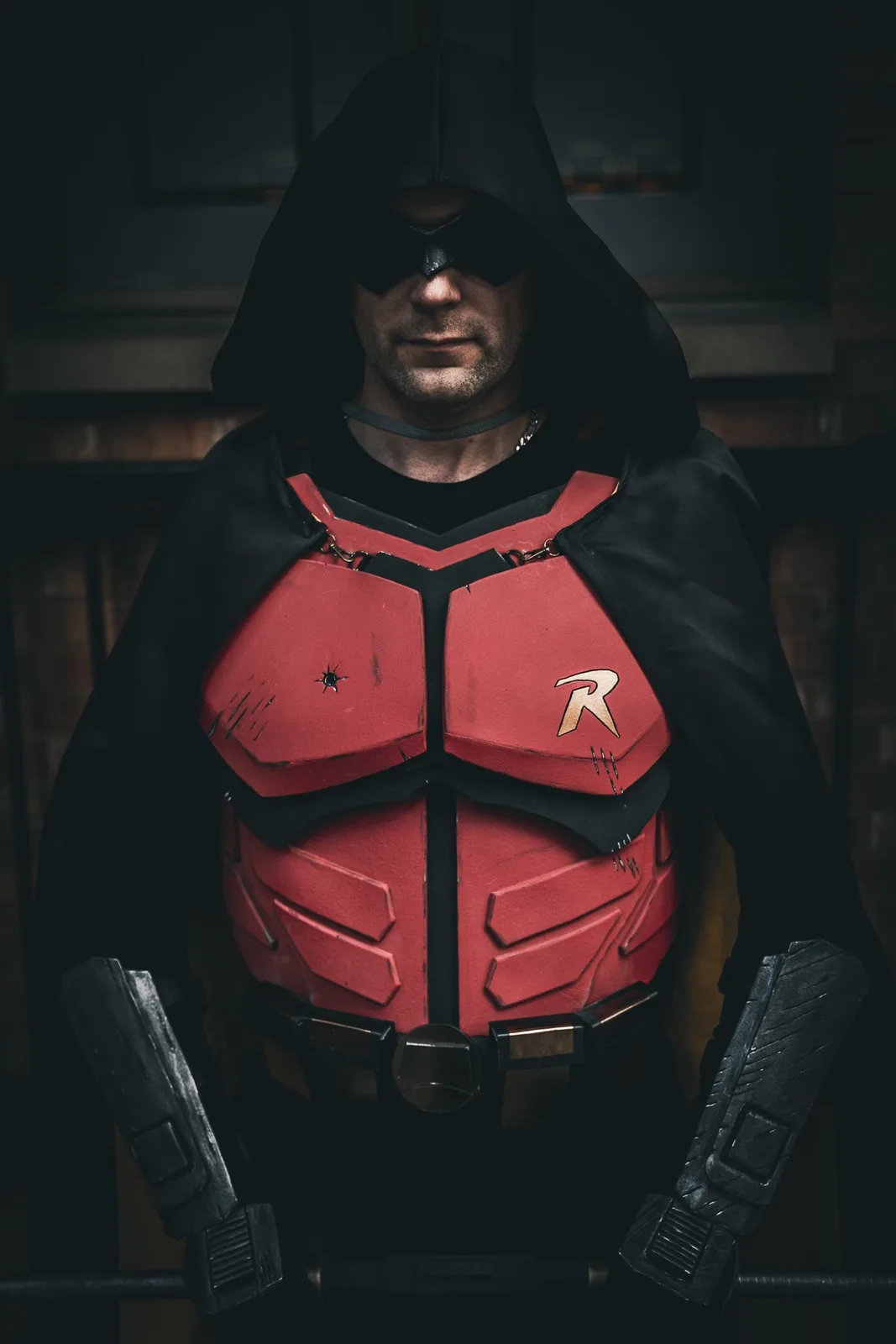 A person in a dark, hooded costume resembling the superhero Robin, with a red armored chest piece featuring a stylized 'R'…
