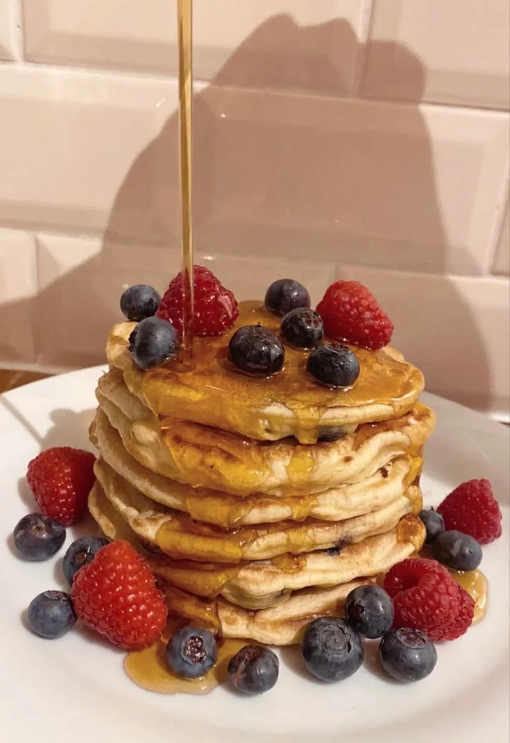 A tall stack of golden-brown pancakes on a white plate, topped with fresh blueberries and raspberries, with syrup being…