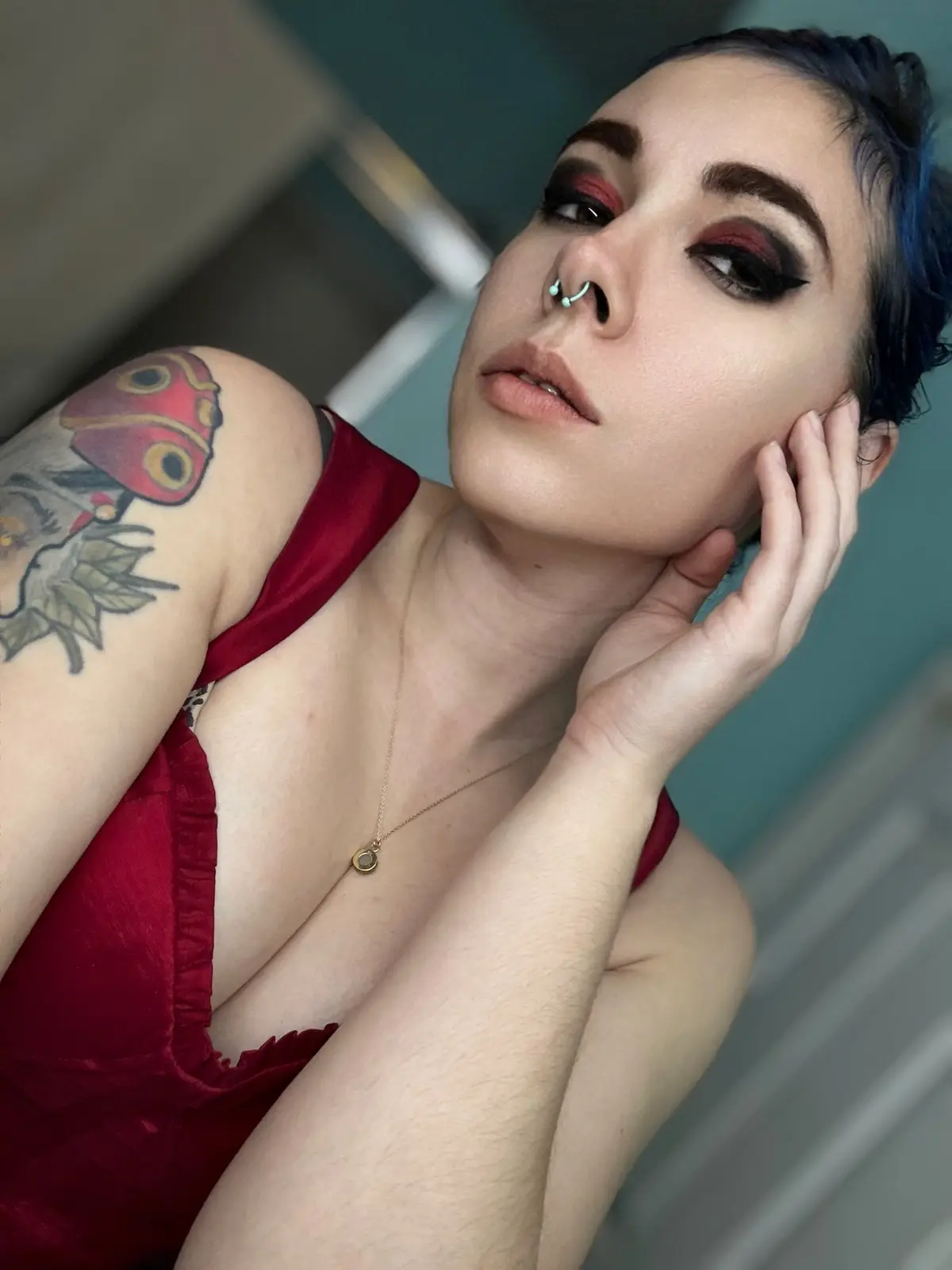 A person with blue-dyed hair, heavy red and black eye makeup, and a nose ring poses in a red top, resting their chin on…