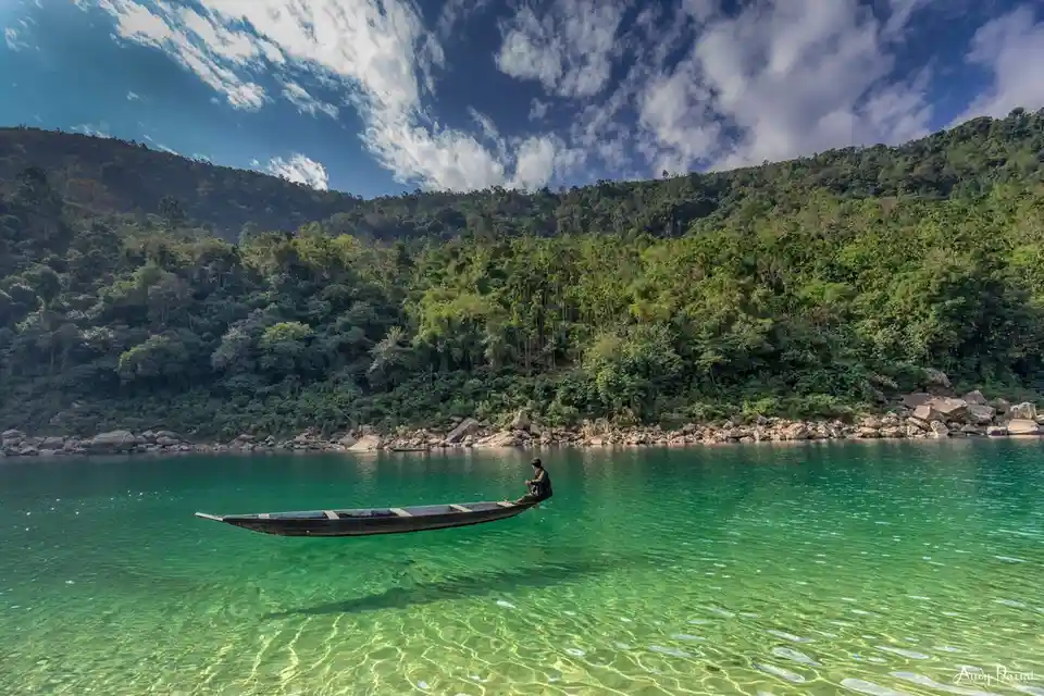 A person sits alone in a small wooden boat on a calm, emerald-green body of water, surrounded by lush green hills and a…