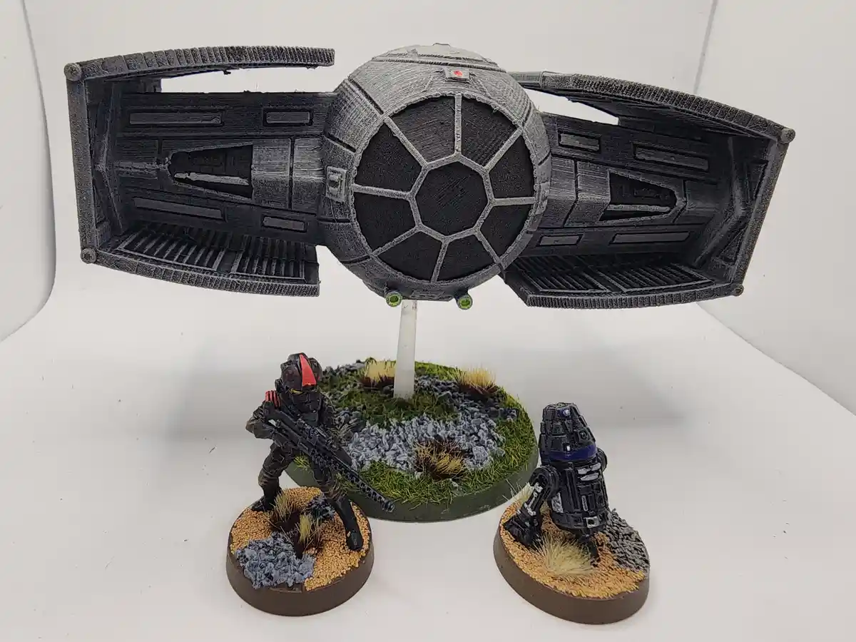 A 3D-printed model of a TIE fighter is mounted on a stand, hovering above a miniature diorama with a painted human-like…