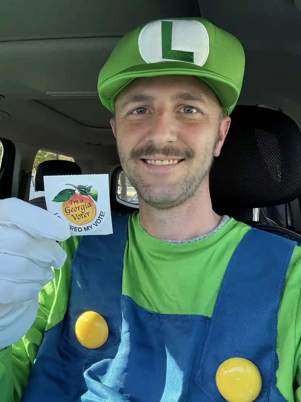 A man dressed as Luigi from Super Mario, wearing a green cap with an 'L', blue overalls with yellow buttons, and white…