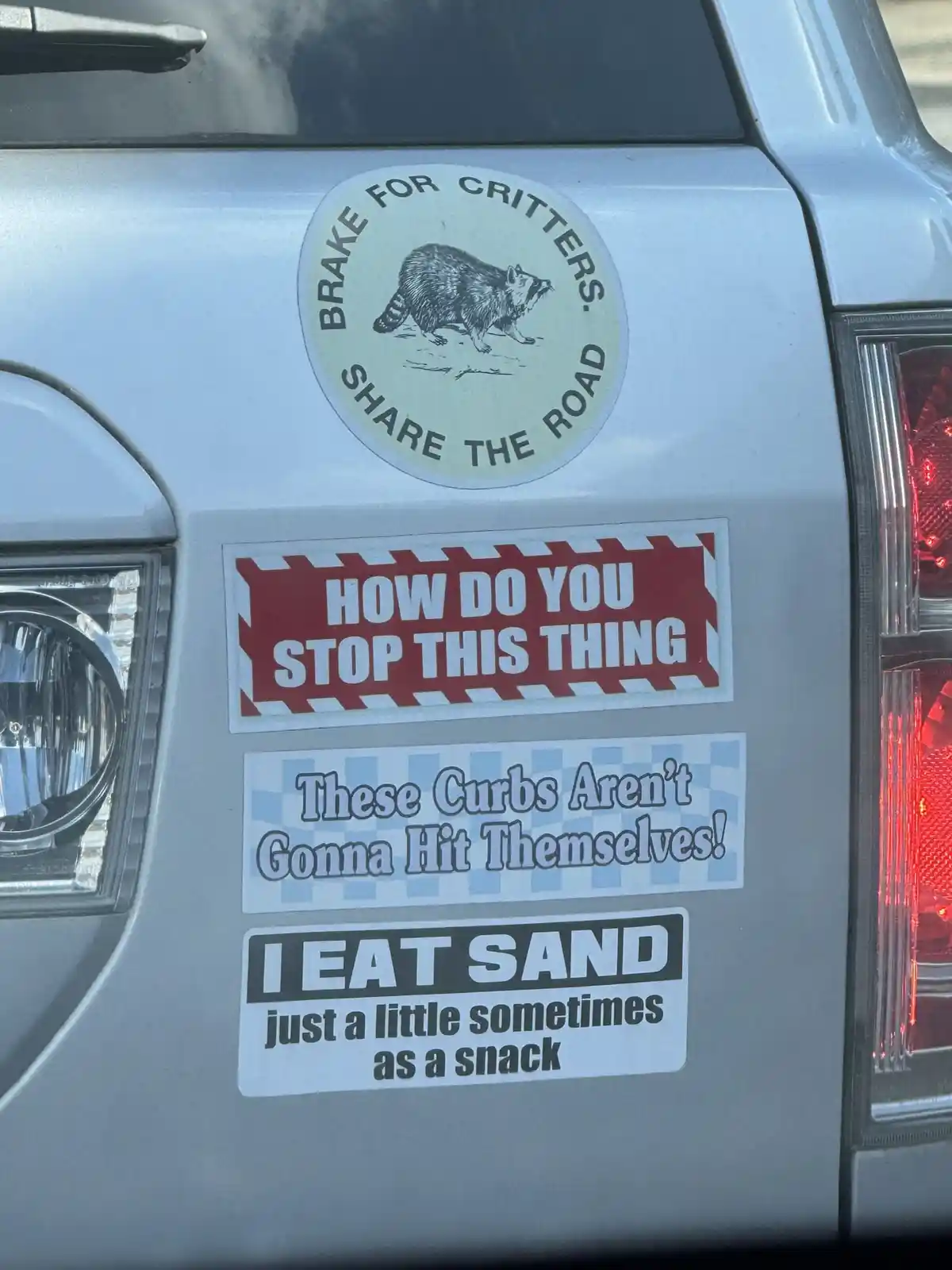 A close-up of a silver vehicle's rear door shows four humorous bumper stickers. The top circular sticker features a raccoon…