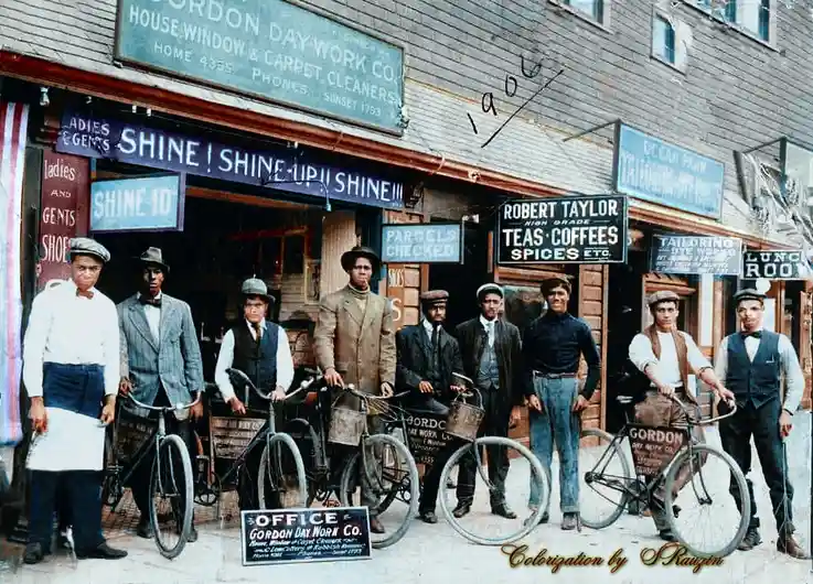A colorized historical photo shows eight men posing with bicycles in front of a row of early 20th-century storefronts with…