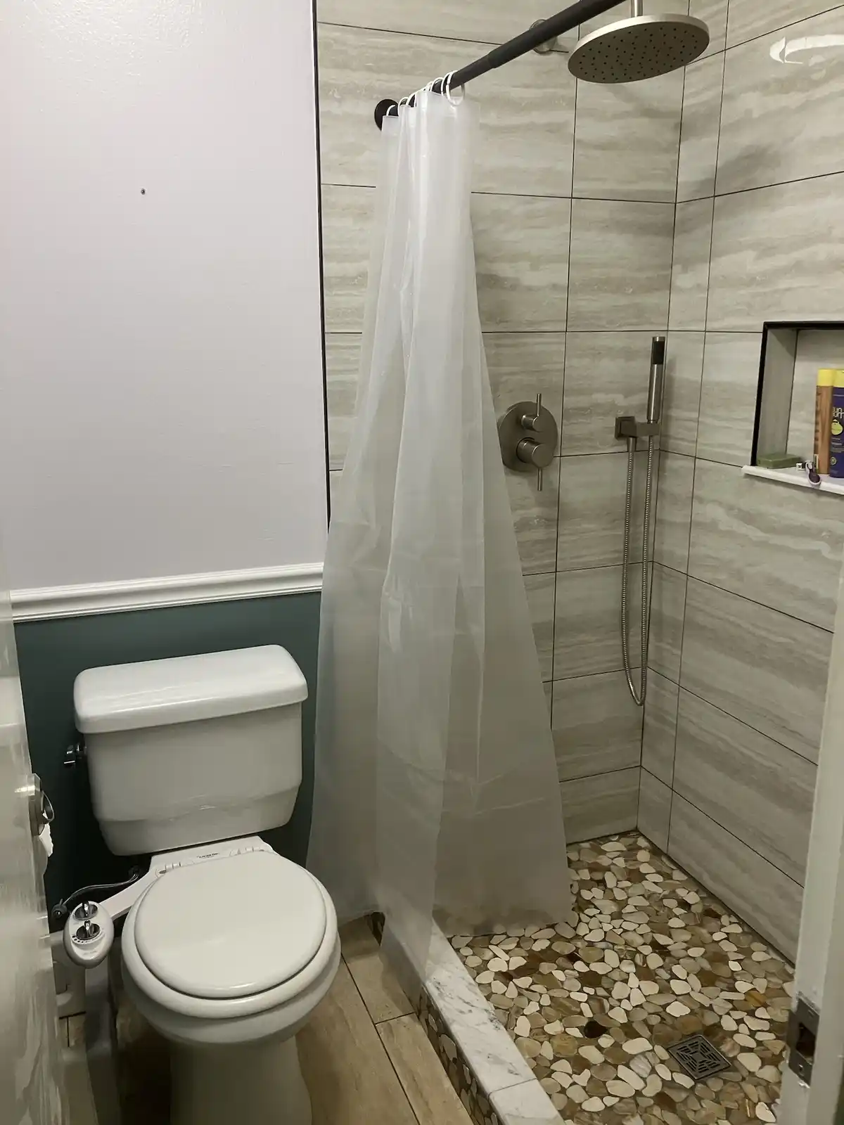 A bathroom photo shows a white toilet next to a walk-in shower with a translucent white shower curtain. The shower has gray…