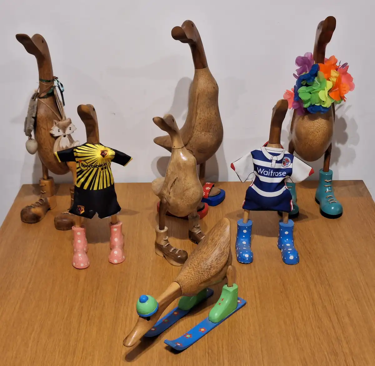 Several wooden duck figurines are arranged on a wooden table, each dressed in different outfits including sports jerseys…