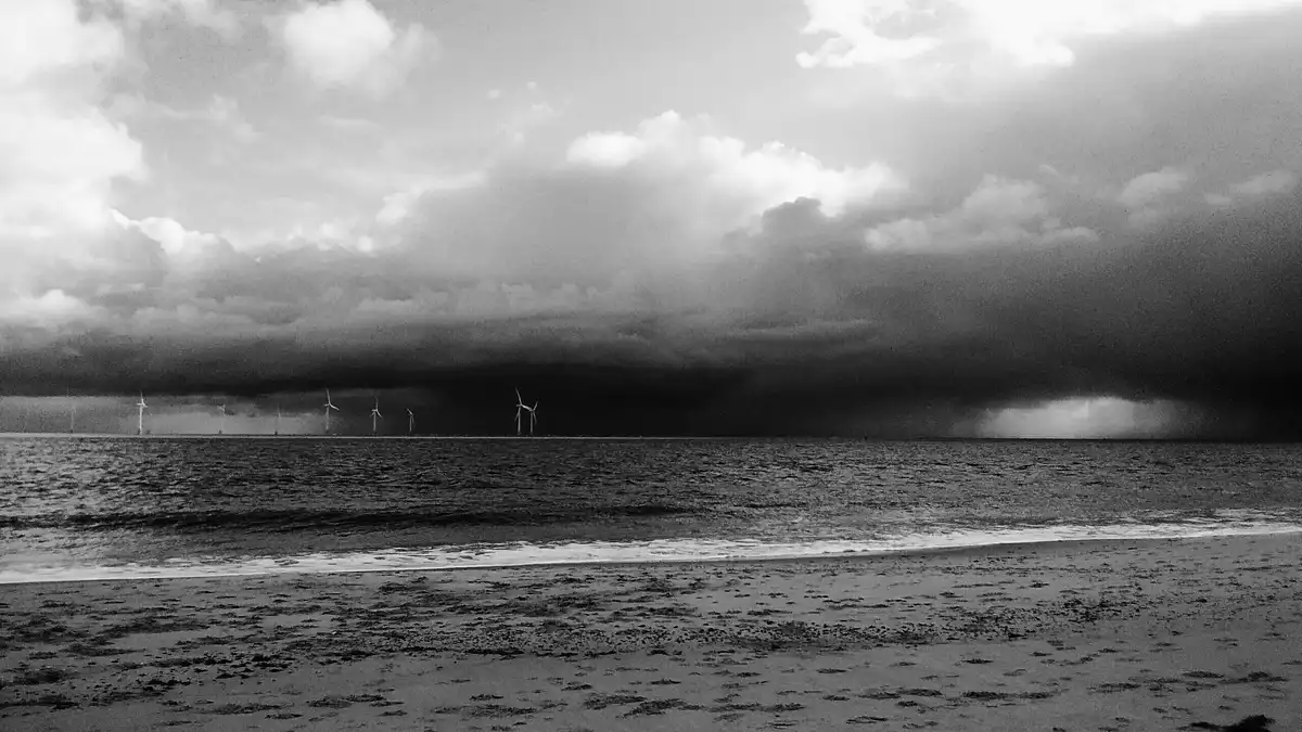 A black and white photo of a beach with waves lapping the shore, a dark storm cloud bank looming over the horizon, and…