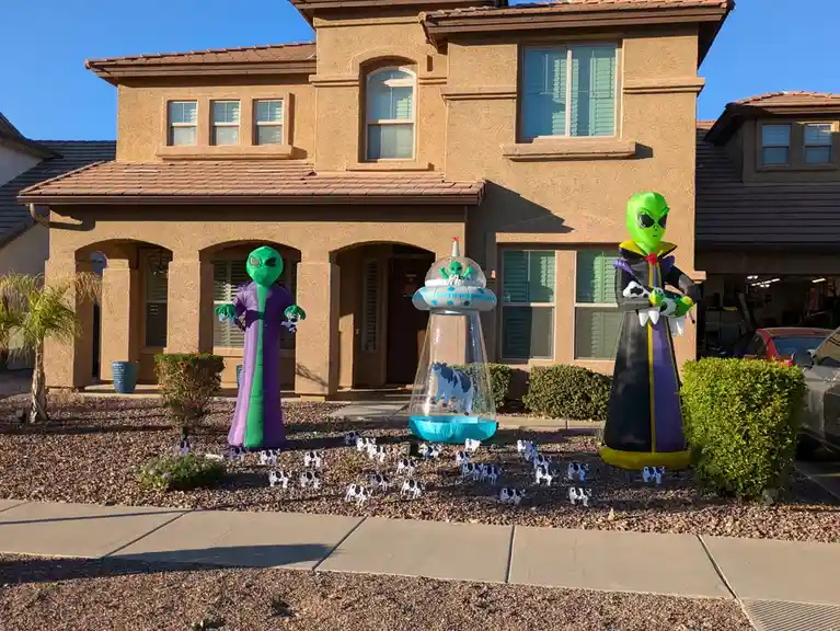 A suburban house's front yard is decorated with large inflatable alien figures and a UFO containing a cow, surrounded by…
