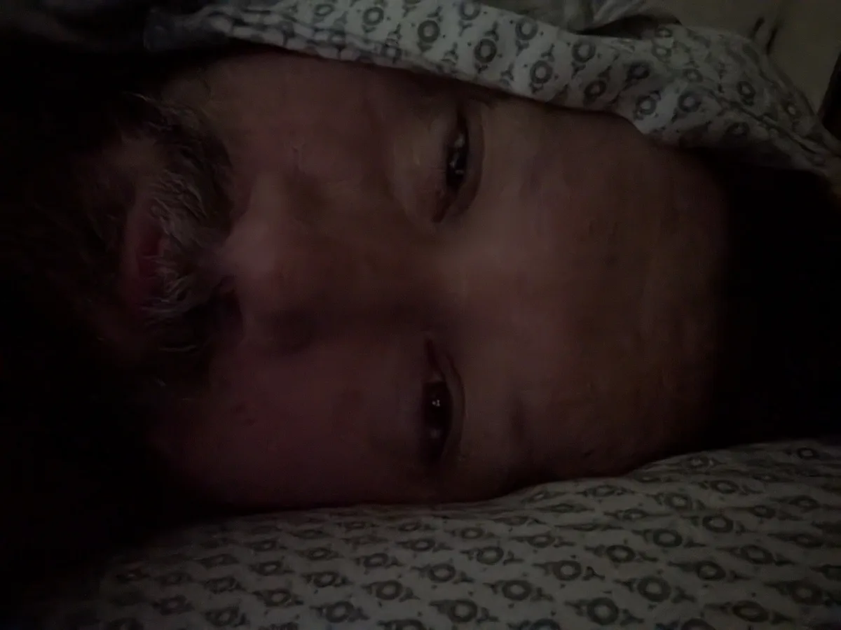 A dimly lit close-up of a person lying in bed, eyes open and staring, partially covered by a patterned blanket. The overlaid…