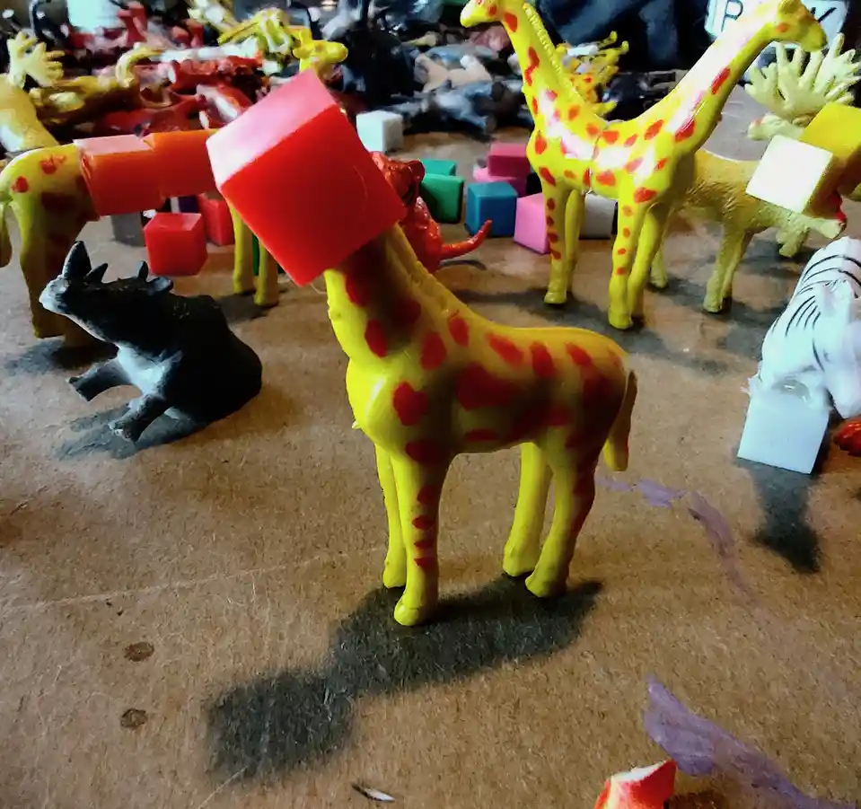 A yellow plastic giraffe toy with red spots has a large red cube balanced on its head, surrounded by other plastic animal…