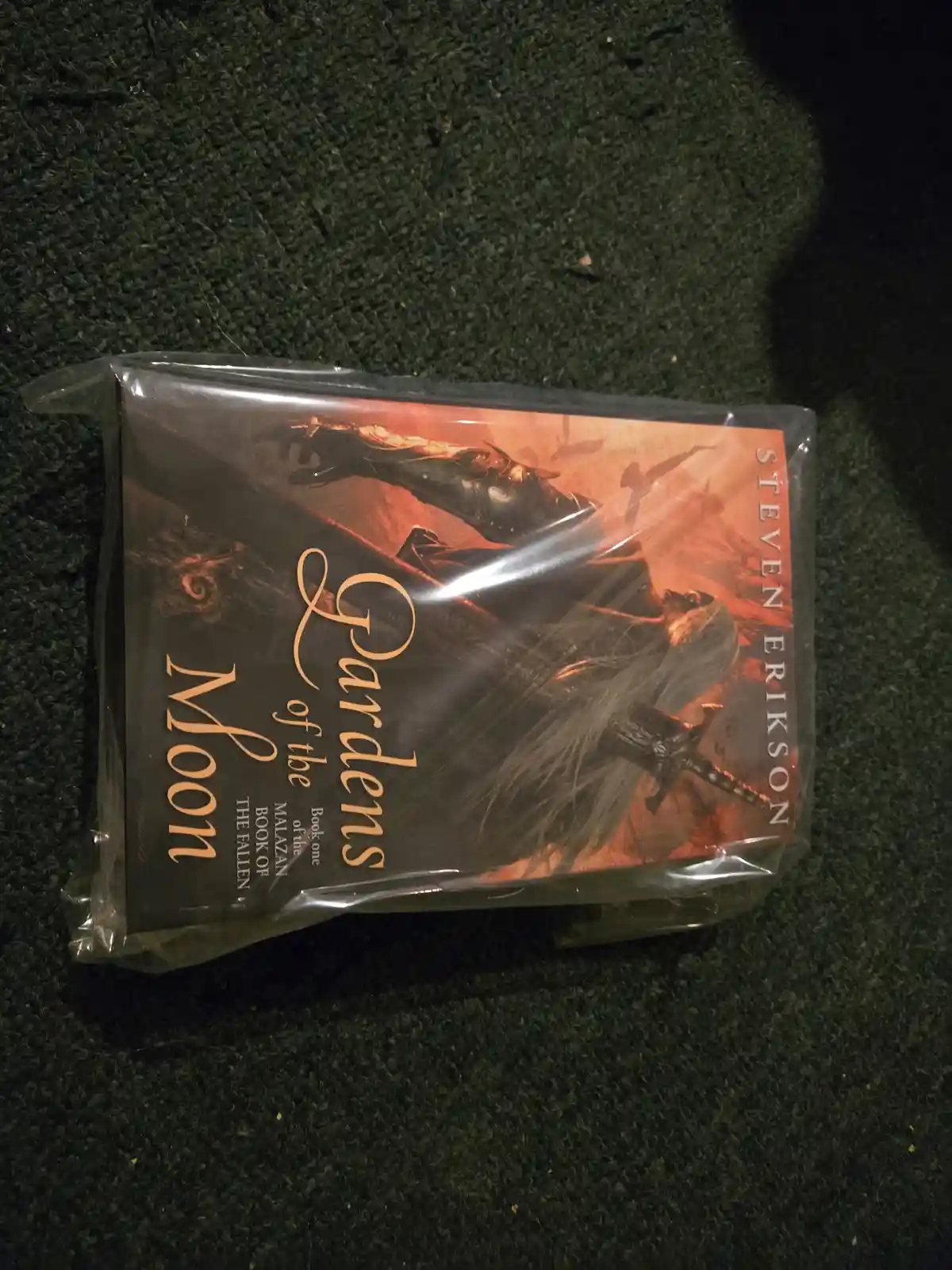 A book titled 'Gardens of the Moon' by Steven Erikson, still sealed in plastic wrap, rests on a dark green carpet. The cover…